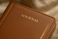 A luxurious close-up of a vegan-leather journal cover with visible stitching and embossed foil detailing. The shot emphasizes texture and craftsmanship. Background is blurred linen fabric with warm neutral tones. Lighting is soft but directional to highlight quality.